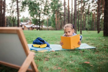 Okula geri döndüm. Salgın gerçekliğinde açık hava dersi sırasında yeşil çimlerin üzerinde oturan ilkokul öğrencisi kız. Güvenli melez eğitimi, sosyal mesafe kuralı, yeni okul rehberliği. 