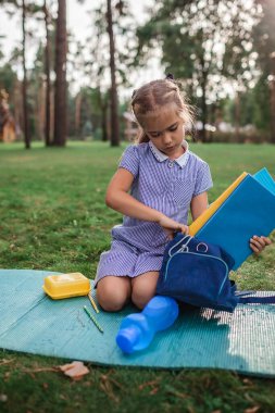 Okula geri döndüm. İlkokul öğrencisi kız otların üzerinde oturuyor ve salgın gerçekliğinde açık hava dersinden sonra kitap paketliyor. Güvenli melez eğitimi, sosyal mesafe kuralı, yeni okul rehberliği. 