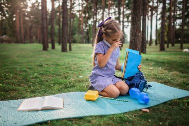 Okula geri döndüm. İlkokul öğrencisi kız, salgın gerçekliğinde açık hava dersinde çimenlerde oturup burnunu siliyor. Güvenli melez eğitimi, sosyal mesafe kuralı, yeni okul rehberliği. 