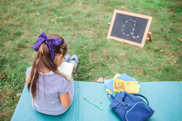New normal back to school. Elementary scholar girl sitting on the green grass during open-air class in pandemic reality. Safe hybrid education, social distance rule, new schooling guidance