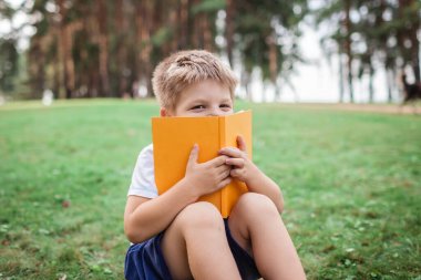 Okula geri döndüm. Anaokulu öğrencisi, salgın sırasında elinde kitapla yeşil çimlerde oturuyor. Güvenli melez eğitimi, sosyal mesafe kuralı, yeni okul rehberliği. 