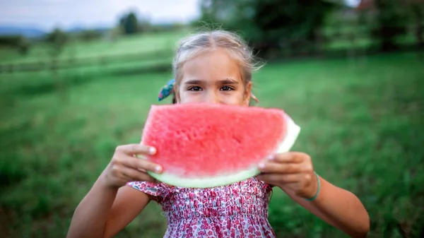 Healthy summer food. Cute kid biting a slice of watermelon in the backyard of farmhouse, happy summertime, countryside, outdoor lifestyle