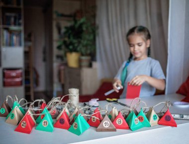 Sevimli küçük çocuk renkli kağıt üçgenli el yapımı takvim yapıyor. Şekerler ve şekerler dalda asılı renkli üçgenlerde saklıdır. Çocuklar için mevsimlik etkinlik, aile kış tatili