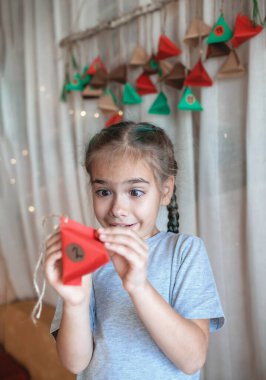 Sevimli küçük çocuk renkli kağıt üçgenli el yapımı takvim açıyor. Şekerler ve şekerler dalda asılı renkli üçgenlerde saklıdır. Çocuklar için mevsimlik etkinlik, aile kış tatili