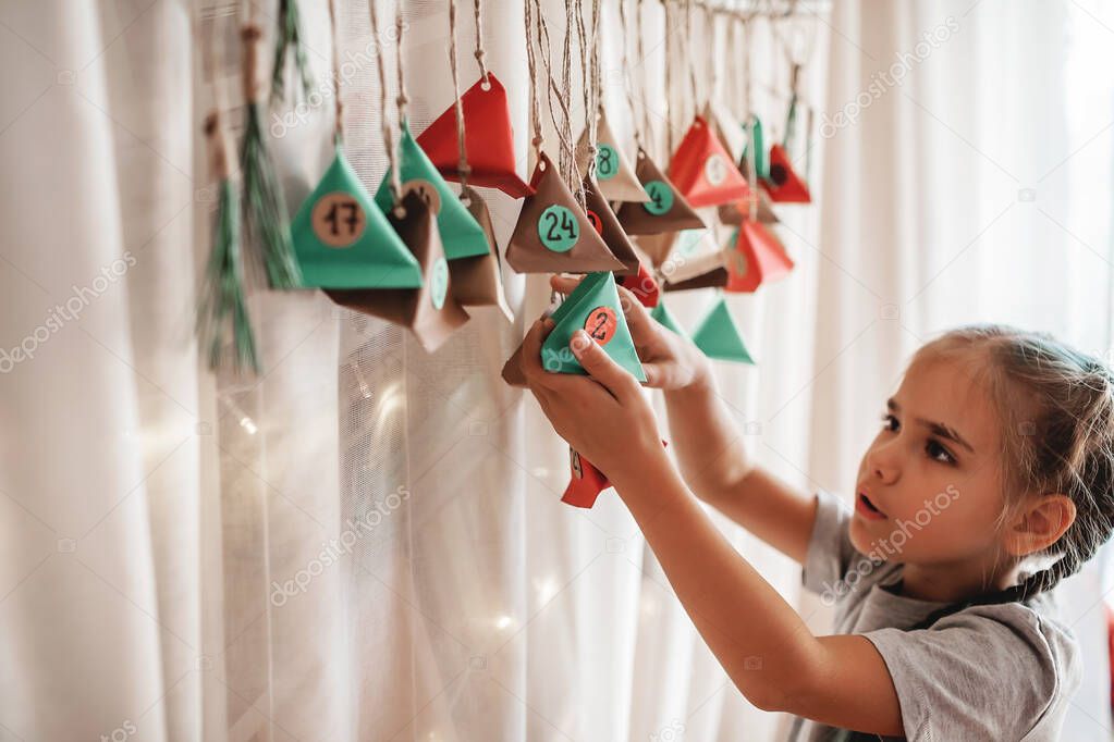 Cute little kid opening handmade advent calendar with color paper triangles. Sweets and candies are hidden in colorful triangles hanging on branch. Seasonal activity for kids, family winter holidays
