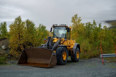 Sarı ön yükleyici traktör, sonbahar ağaçlarıyla çakıl taşlarının üzerine park edilmiş.. 