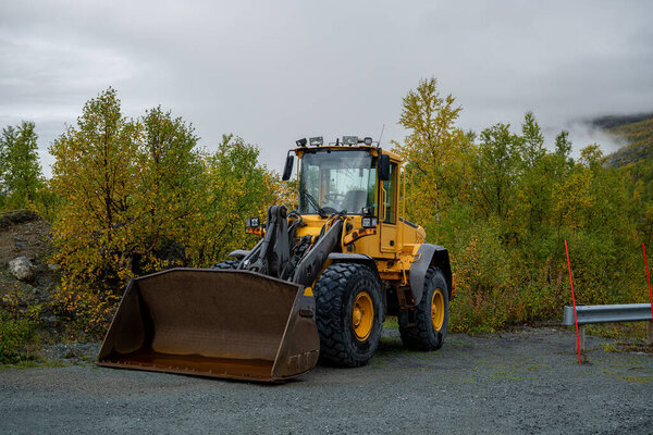 Желтый передний погрузчик припаркован на открытом воздухе на гравии с осенними деревьями. 