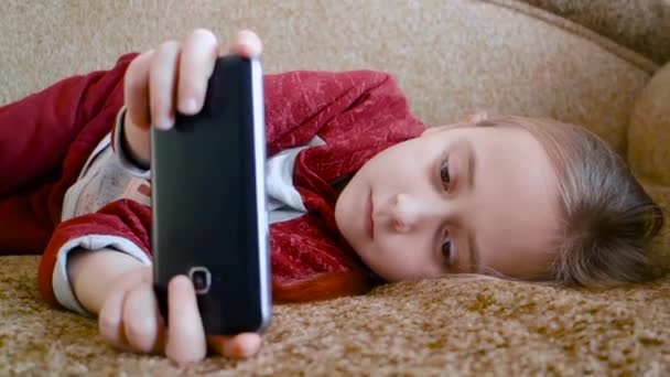 Дівчина-підліток дивиться відео в телефоні, лежачи на дивані . — стокове відео