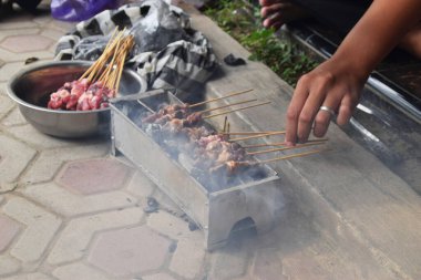 A man's hands grilling goat satay over charcoal on a grill. Grilled goat satay is often served during Eid al-Adha celebrations in Indonesia.