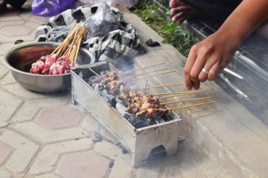 A man's hands grilling goat satay over charcoal on a grill. Grilled goat satay is often served during Eid al-Adha celebrations in Indonesia.