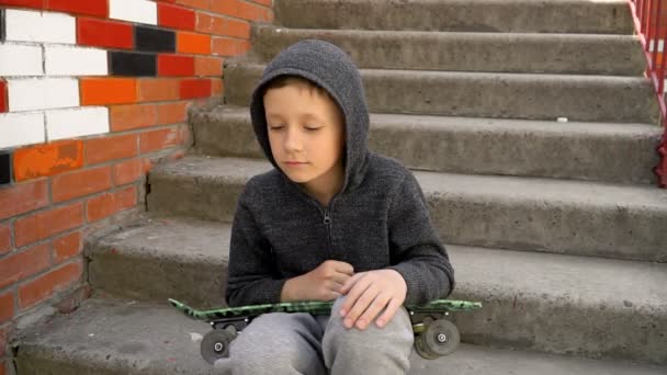 Мальчик сидит на ступеньках и держит скейтборд в руках. — стоковое видео