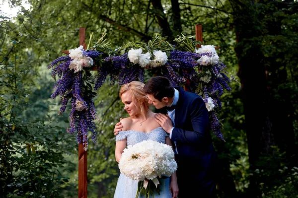 The bride and groom stand at the arch, decorated with flowers, with a large bouquet of white peonies.On-site registration in the Park Wedding walk and photo shoot