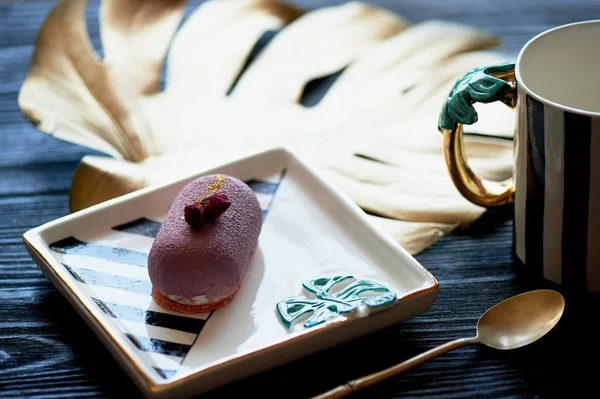 Purple fruit mousse cake, on a beautiful saucer.On a dark background, with a sheet of monstera painted in gold paint. Culinary art