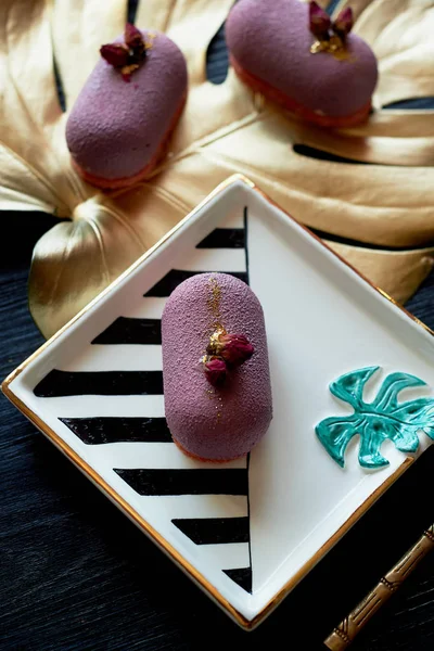 Purple fruit mousse cake, on a beautiful saucer.On a dark background, with a sheet of monstera painted in gold paint. Culinary art