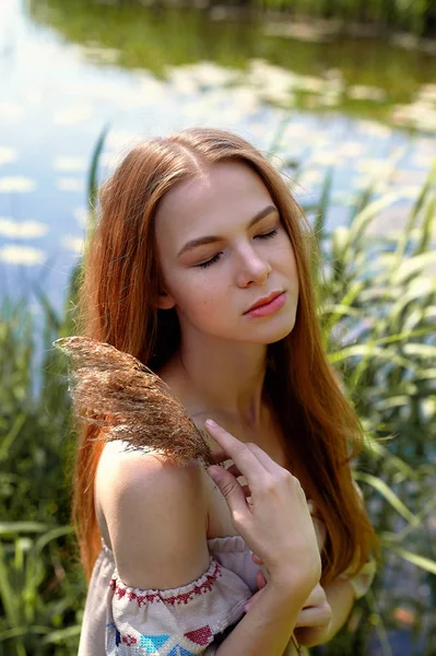 Portret van een roodharige meisje met lang haar. Haar neer. Artistieke portret. Kind van aard. Nabijheid van natuurlijkheid en natuur. Een eenvoudige linnen jurk. In de achtergrond, een rivier- of meerwater — Stockfoto