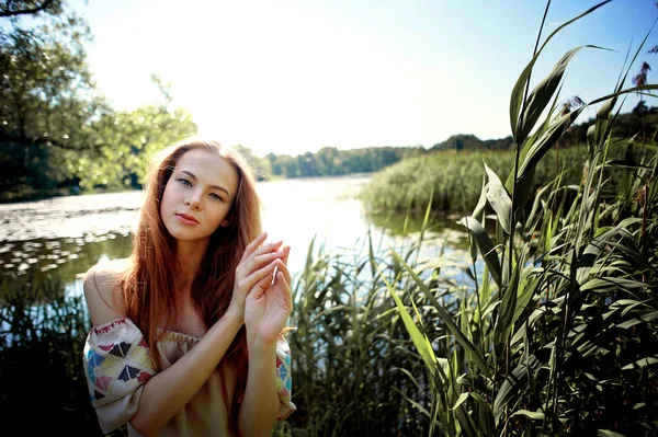 Portret van een roodharige meisje met lang haar. Haar neer. Artistieke portret. Kind van aard. Nabijheid van natuurlijkheid en natuur. Een eenvoudige linnen jurk. In de achtergrond, een rivier- of meerwater — Stockfoto