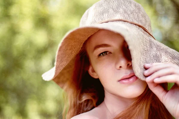 A sophisticated red-haired girl in a simple linen dress, in a light wide-brimmed hat.Model look. Natural beauty. Artistic portrait. Keeps the brim