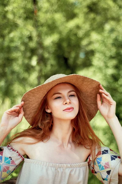 A sophisticated red-haired girl in a simple linen dress, in a light wide-brimmed hat.Model look. Natural beauty. Artistic portrait. Keeps the brim