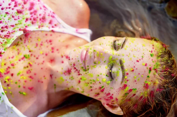 Festival Holi.The radost a štěstí. Malování na obličej. Blond holka. Spočívá v čisté vodě na oblázkové pláži — Stock fotografie