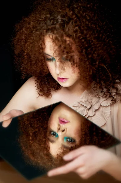 Девушка с пышными вьющимися рыжими волосами. Глядя в зеркало. Тайна, предчувствие будущего. Его отражение. Загляни внутрь себя. Самопознание — стоковое фото