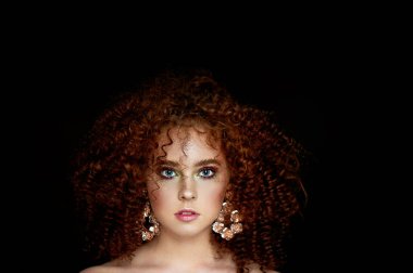 Yemyeşil kıvırcık kırmızı saçlı bir kız. Yüzü parlak makyaj ve büyük küpe ile kapatın. Mor gölgeler.