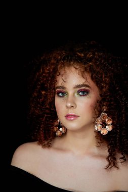 Yemyeşil kıvırcık kırmızı saçlı bir kız. Yüzü parlak makyaj ve büyük küpe ile kapatın. Mor gölgeler. Çıplak omuz.