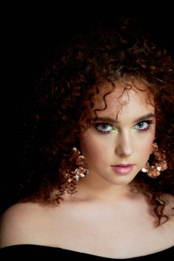 Yemyeşil kıvırcık kırmızı saçlı bir kız. Yüzü parlak makyaj ve büyük küpe ile kapatın. Mor gölgeler. Çıplak omuz.