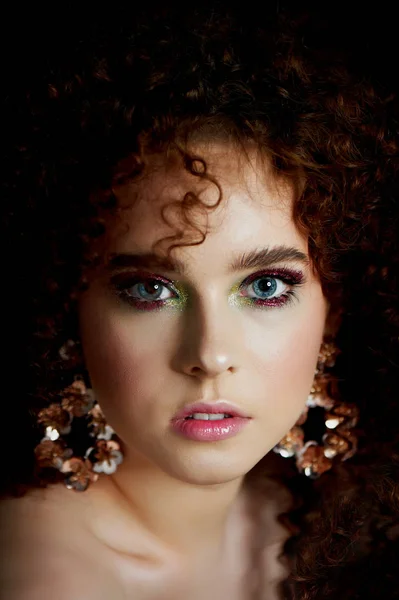 Una chica con pelo rojo rizado exuberante. Cara cerrada con maquillaje brillante y pendientes masivos. Sombras púrpuras . —  Fotos de Stock