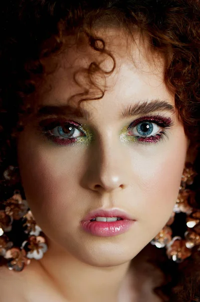 Yemyeşil kıvırcık kırmızı saçlı bir kız. Yüzü parlak makyaj ve büyük küpe ile kapatın. Mor gölgeler.