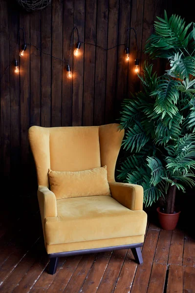 Acogedor sillón de color mostaza, palmera y paredes de madera. Interior en estilo retro. Tibia guirnalda amarilla luz  . — Foto de Stock