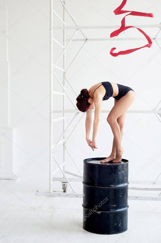 Gymnast in a black swimsuit is on a black barrel.On light background.Red ribbon.