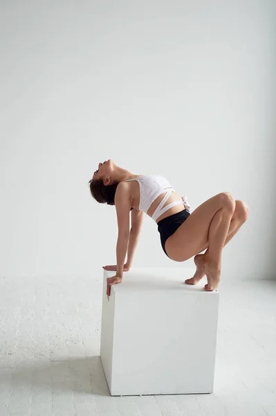 Прекрасная балетная танцовщица позирует в студии, исполняя отклонение в спине и опираясь на большой куб — стоковое фото