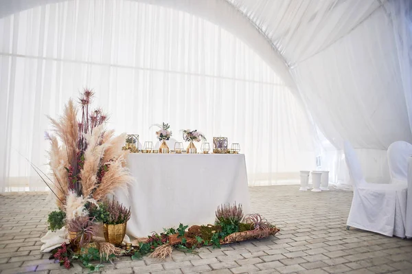 Boho tarzında düğün dekorasyonu. Açık renkler, çadırda. Pampas çimile dekore düğün masası. Güller ve altın geometrik şamdanlar.