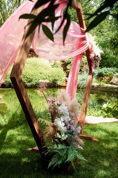 Enregistrement dans la forêt au bord du lac. Une arche de bois en forme de diamant décorée de pampas, mousseline rose. Mariage floristique. — Photo