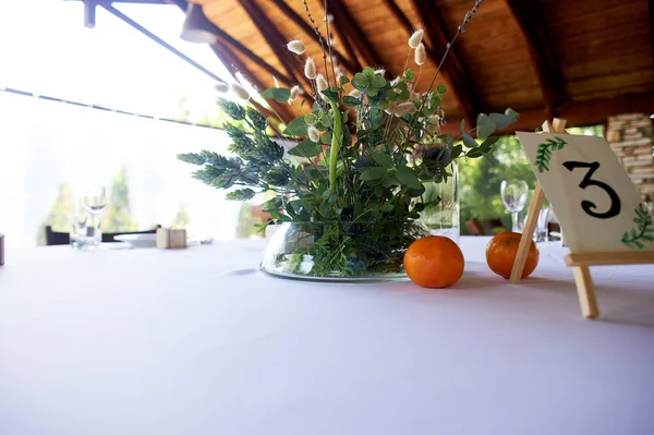 Összetétele szántóföldi gyógynövények és gabonafélék az asztalon az étteremben egy átlátszó vázában.Fehér terítő és két mandarin. Halandóság. — Stock Fotó