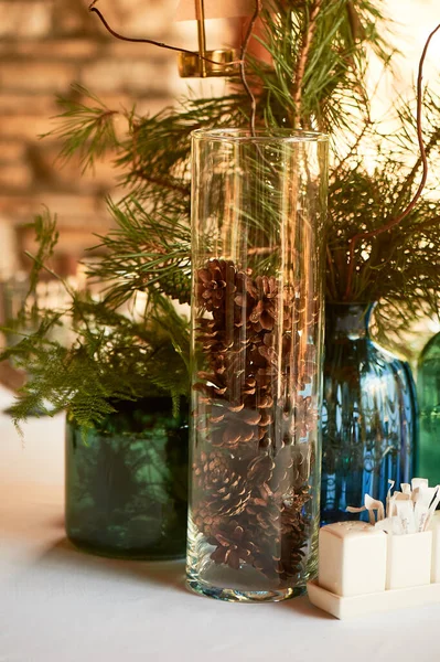 Tischdecken im Restaurant, dekorative Floristik. Flaschen aus blauem und grünem Glas mit Fichtenpfoten. Die Kegel im transparenten Gefäß. Öko-Stil. — Stockfoto