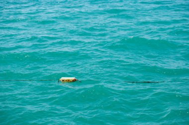 Tek, dikdörtgen beyaz ve sarı bir şamandıra, koyu renkli bir kabloya bağlı, deniz mavisi bir okyanusta yüzer..