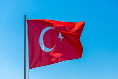 Bir Türk bayrağı açık mavi gökyüzüne doğru dalgalanıyor. Beyaz hilal ve yıldızlı bayrak ağırlıklı olarak kırmızıdır..