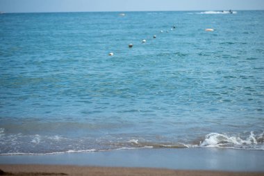 Sakin bir deniz manzarasında kumlu bir sahil buluşması, açık mavi bir su, mesafeye kadar uzanan bir şamandıra ve görülebilir su araçları bulunur..