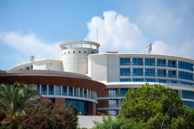 Çok katlı bir binanın dış görüntüsü yuvarlak mimari özellikleri, mavi pencereleri ve mavi gökyüzünün altında çevreleyen yeşillik..