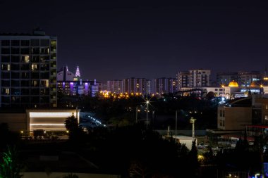 Aydınlatılmış binalar ve karanlık ağaçlarla dolu bir şehir manzarası. Çeşitli mimari stiller ve çeşitli ışık kaynakları görülebilir.