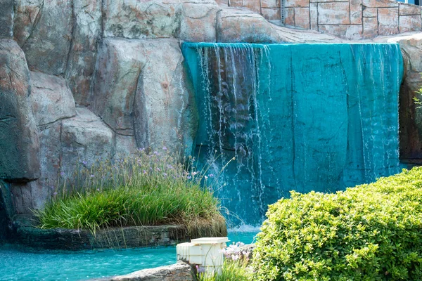 Mavi renkli bir şelale, temelinde ve çevresinde canlı yeşil bitkiler bulunan yapay bir kaya oluşumunun üzerine dökülür..