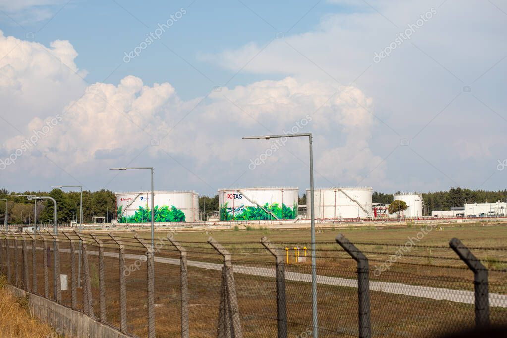 View of three white industrial storage tanks with vegetation-themed graphics, seen behind a chain-link fence and tall, grey lighting poles.