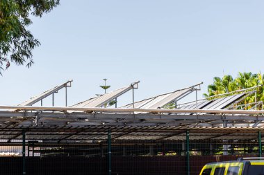 Çerçevenin kenarlarında yeşillik olan mavi gökyüzüne karşı metal bir çerçeve üzerine monte edilmiş açılı güneş panellerinin görüntüsü.