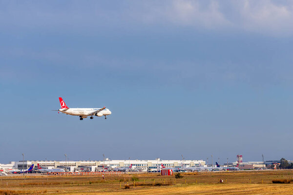 A Turkish Airlines plane approaches for landing at an airport with other aircraft visible at terminals in the background.