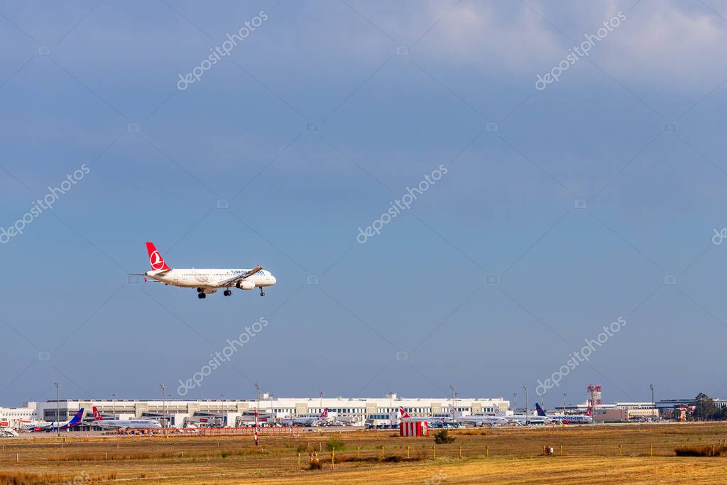 A Turkish Airlines plane approaches for landing at an airport with other aircraft visible at terminals in the background.