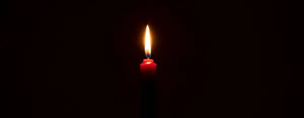 Single burning candle flame or light is glowing on a small red candle on black or dark background on table in church for Christmas, funeral or memorial service with copy space.