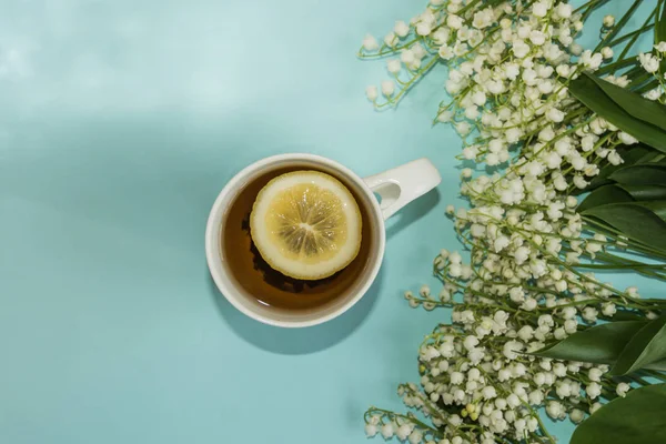 Top view of the cup of tea with the slice of lemon and lily of the valley flowers on the pastel blue background.