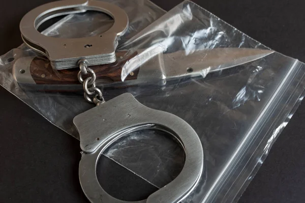 Knife in a plastic package and handcuffs on black color background table. — Stock Photo, Image