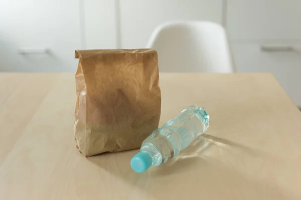 Brown paper lunch bag and bottle of water on wooden table over kitchen background.